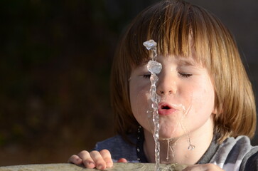 Cute little boy drinking from a fountain. Summer thirst. A baby with long hair catches a jet of...
