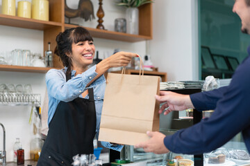Beautiful female cafe owner in apron smiling and giving takeaway food paper bag to customer at coffee shop