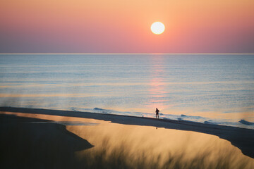 Jogging o zachodzie słońca na morskiej plaży. W tle sylwetka biegacza podczas wieczornego treningu. Kolory zachodzącego słońca, błękit, różowy, pomarańcz, czerwony, fioletowy. Odbicie słońca w wodzie. - obrazy, fototapety, plakaty