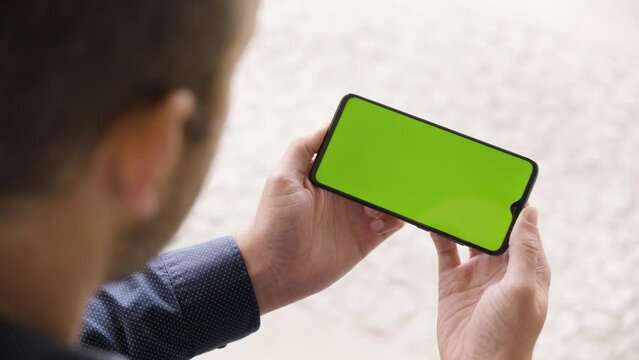 A man looks at a smartphone (horizontal) with green screen - closeup from behind the shoulder