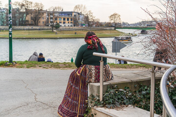 A gypsy woman in national dress rests by the river on the embankment in Krakow on a cloudy day - obrazy, fototapety, plakaty