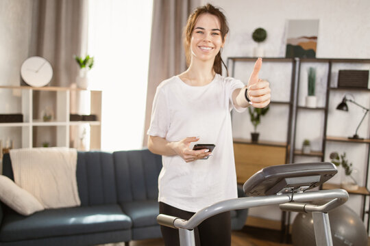 Front view image of a sporty woman using a smartphone while doing an exercise on the treadmill in black sports leggings and a white T-shirt trainig at home showing thumb up.