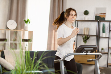 Young beautiful woman running on a treadmill at home. Female runner doing cardio exercise healthily lifestyle. Sporty slender girl sweating workout in modern apartment during leisure activity.