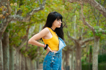 Young girl dressed in yellow and wearing a denim overalls in profile with a park background full of trees in autumn shades of green and yellow