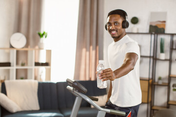 Way to health. Handsome african american man athlete preparing for cardio training on treadmill and listening to music with headphones looking at the camera at home, free space.