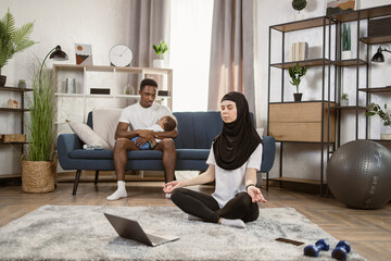 Arabian muslim woman in hijab meditating on mat in the living room using laptop as a tutorial for...