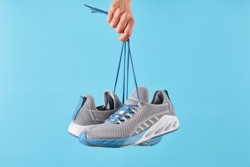 Hand holds hanging gray running sneakers by the laces on blue pastel background. Hand with a new...