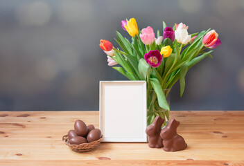 Easter concept. Chocolate Easter eggs in wicker basket, chocolate bunny, tulip flowers and white frame on wooden table.
