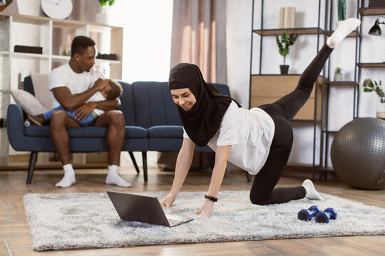 Fit muslim arabic woman doing yoga plank and watching online tutorials on laptop while training in living room and her african husband puts little son to sleep while sitting on the couch.