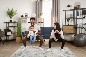 Young multinational multiracial couple doing squats training together at home, african man holding in his arms their cute little son and arab woman working out together standing in living room.