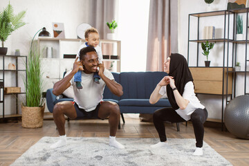 Obraz premium Muscular Athletic african Man holds his son on his shoulders and Beautiful Fitness muslim Woman in Workout Clothes are Doing Squat Exercises in Their Bright and Spacious Apartment.