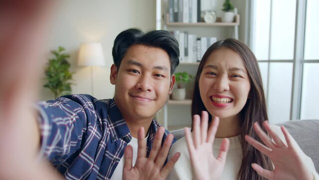 POV happy Asian couple man and woman making video call, selfie, waving to camera via mobile phone talking with friends