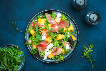 Delicious summer fresh salad with cantaloupe melon, prosciutto, soft cheese and arugula on blue table background, top view, copy space