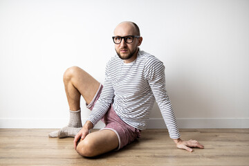 Serious young man in glasses sitting on a wooden floor against a white wall.
