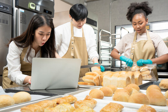 Selective focus of three young bakers, Asian man and woman, African woman, standing at table using laptop, slicing bread, and packing bread into bag, prepare to deliver breads according to the order.