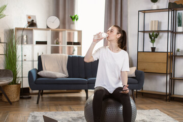 Young caucasian woman in black sports leggings and a white t-shirt with smart phone sitting on fit ball drinking water on the background of bright modern apartment. Sport workoutat at home.
