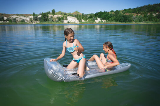 Two little cute girls in swimsuits swim on an inflatable mattress on a blue career lake.