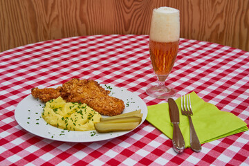 Wiener snitzel served on the white plate with mashed potatoes, melted butter, pickled sour cucumbers and chopped chives together with the glass of pilsener bier with green napkin and fork and knife.