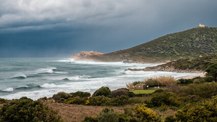 Rough seas crash onto the beach at Ghjunchitu in the Balagne region of Corsica with the lighthouse of Ile Rousse in the distance