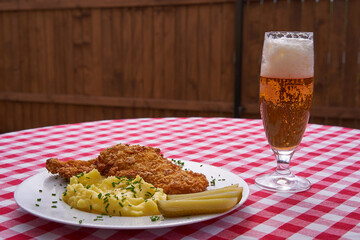 Wiener schnitzel with mashed potatoes, melted butter, chopped chives and pickled cucumer on the rustic white plate. Served on the table in garden restaurant together with czech style pilsner beer.