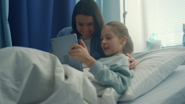 Smiling mom visiting daughter play on tablet computer together in hospital room
