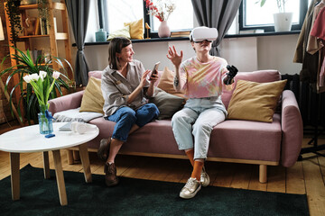Girl with prosthetic arm in goggles playing virtual reality game with her friend while she using mobile phone on sofa in living room