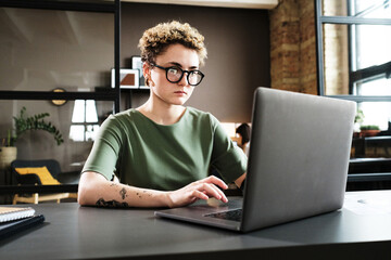 Young woman in eyeglasses concentrating on her online work on laptop at her workplace at office