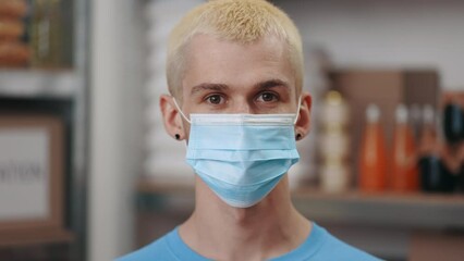 Portrait of caucasian young man in medical face mask standing among foodbank warehouse. Blue background. Donation, pandemic and people concept. - Powered by Adobe