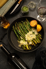 Fried green asparagus with lemon and parmesan in a pan.  Set table with plates,cutlery,olive oil,pesto and bbottle of  wine.  Dark background. Top view.