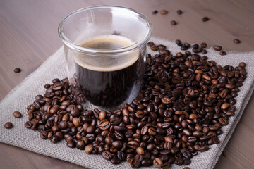 Close-up of glass cup with coffee and pile of roasted coffee beans on piece of burlap on wooden surface. Heat-resistant coffee glass with double walls on table. Selective focus.
