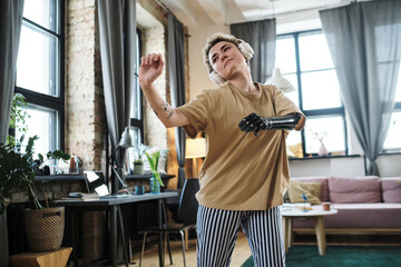 Young woman with prosthetic arm listening to music in wireless headphones and learning to dance in living room
