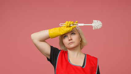 tired maid with a toilet brush in her hand wipes sweat from her forehead on a pink background