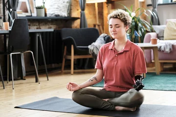 Rolgordijnen Girl with prosthetic arm sitting in lotus position and doing yoga in living room © AnnaStills