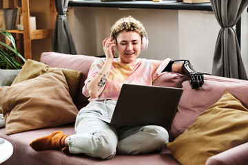Happy young woman with prosthetic arm wearing wireless headphones talking online using her laptop...