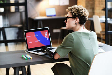 Rear view of young designer with prosthetic arm touching her neck from exhaustion while working with online presentation on laptop