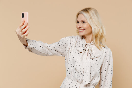 Vivid elderly gray-haired blonde woman lady 40s years old wear pink dress doing selfie shot on mobile cell phone post photo on social network isolated on plain pastel beige background studio portrait