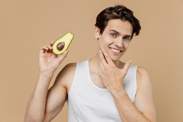 Attractive young man 20s perfect skin wear undershirt hold half of avocado touch face skin isolated...