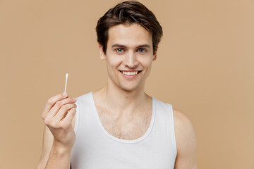 Attractive young man 20s perfect skin in undershirt hold cotton swab stick for ear clean isolated on pastel pastel beige background studio portrait. Skin care healthcare cosmetic procedures concept