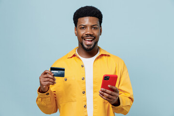 Young fun man of African American ethnicity in yellow shirt using mobile cell phone hold credit...