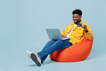Full body young man of African American ethnicity in yellow shirt sit in bag chair use laptop computer credit bank card shopping online order delivery isolated on plain pastel light blue background - Powered by Adobe