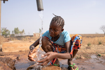 Thirsty African girl refreshes herself with cool drinking water from a village tap; water scarcity...