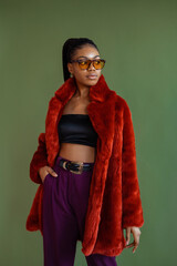 Fashionable confident Black woman wearing trendy orange color faux fur coat, crop top, purple trousers, sunglasses with leopard frame, yellow glass, posing on green background
