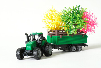Green toy tractor transports green, pink and yellow spring seedlings in a trailer. White background. Copy space. Selective focus.