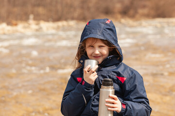Little cute girl sitting on the riverbank, drinking tea from thermos.