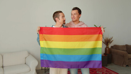 Two young attractive man are standing together, looking at the camera and holding a lgbt flag in...