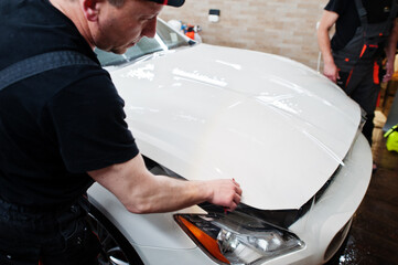 Worker in detailing garage put polyurethane anti-gravel film cover in white luxury car.