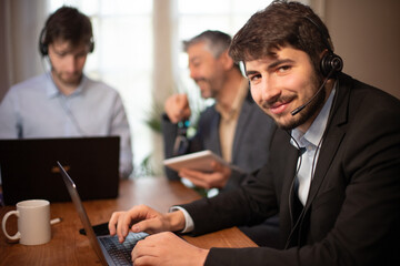 jeune homme d'affaires ou employé de bureau souriant qui travaille au bureau avec ses collègues en arrière plan. il a un micro casque pour participer a une réunion à distance ou répondre au téléphone