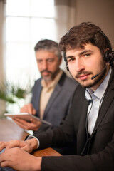 jeune homme d'affaires ou employé de bureau souriant qui travaille au bureau avec ses collègues en arrière plan. il a un micro casque pour participer a une réunion à distance ou répondre au téléphone