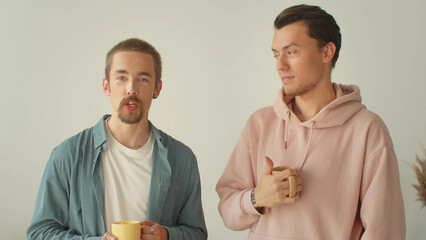 A handsome young man in a pink hoodie is drinking a beverage while his boyfriend is talking to the...