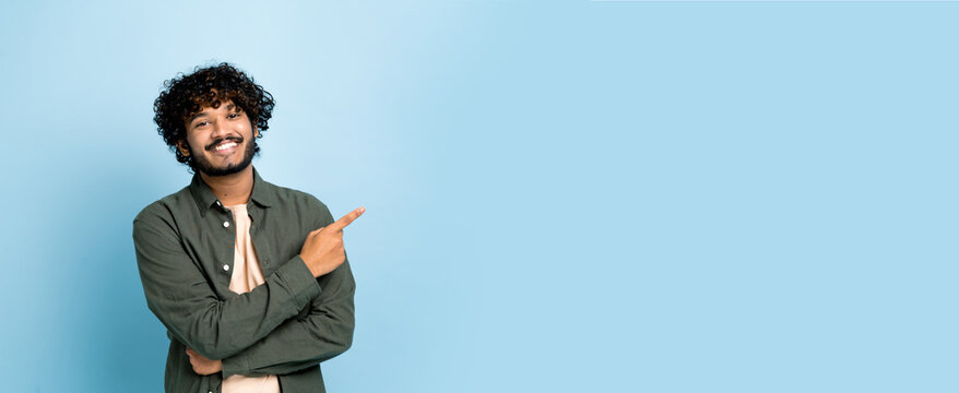 Panoramic photo of arabian or indian guy in casual wear, looks at camera and points finger to the side at empty space for your presentation, stands on isolated blue background, smiling